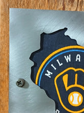 A close-up of a custom metal wall sign featuring the shape of Wisconsin filled with a dark navy blue Milwaukee Brewers logo, mounted on a brushed silver metal plate with a screw visible, set on a wooden surface.